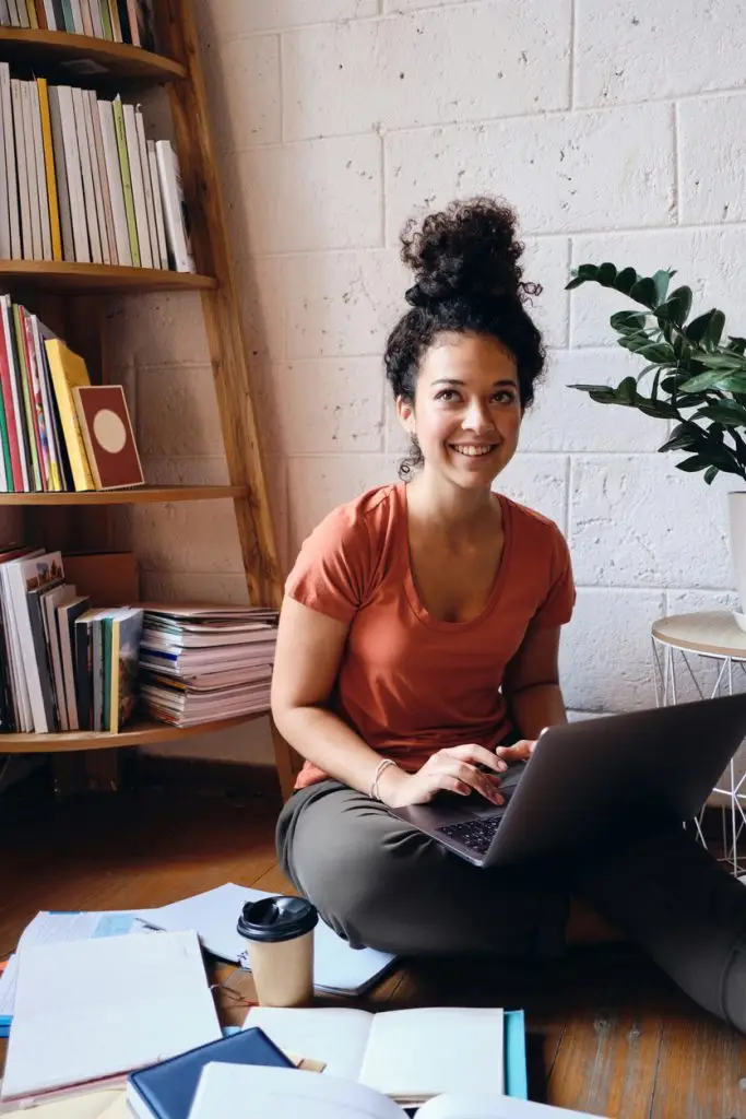 Young beautiful smiling woman with dark curly hair sitting on floor with laptop on knees happily looking aside with books and cup of coffee to go near at cozy home